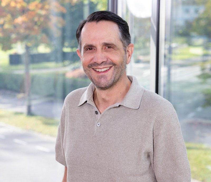 Technischer Mieterservice – Ingo Müller Ein lächelnder Mann mittleren Alters mit kurzem dunklem Haar trägt ein hellbraunes Poloshirt und steht im Freien vor einer grünen, sonnigen Parklandschaft.