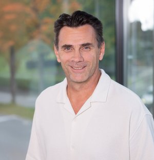 Rickard Merk Projektleiter Porträt eines lächelnden Mannes mittleren Alters mit dunklem Haar, der ein weißes Hemd trägt; im Hintergrund unscharfe Bäume und Fenster.