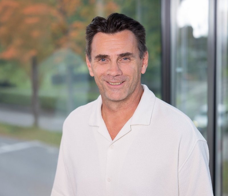 Rickard Merk Projektleiter Porträt eines lächelnden Mannes mittleren Alters mit dunklem Haar, der ein weißes Hemd trägt; im Hintergrund unscharfe Bäume und Fenster.