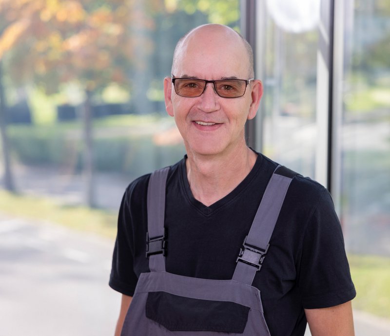 Portrait Ralf Liebitz Porträt eines lächelnden Mannes mittleren Alters mit Brille und Arbeitskleidung, stehend vor einem Fenster mit unscharfem Außenblick.