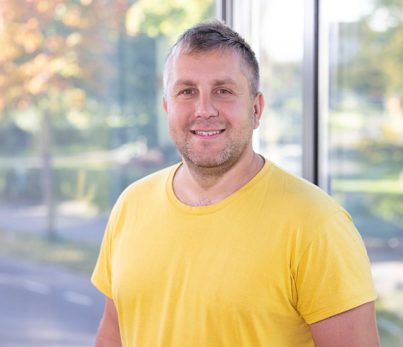 Regiebetrieb – Serhii Kravchenko Porträt eines lächelnden Mannes mit kurzem Haar und Bart, der ein gelbes T-Shirt trägt, vor einem Fenster mit unscharfem Naturhintergrund.