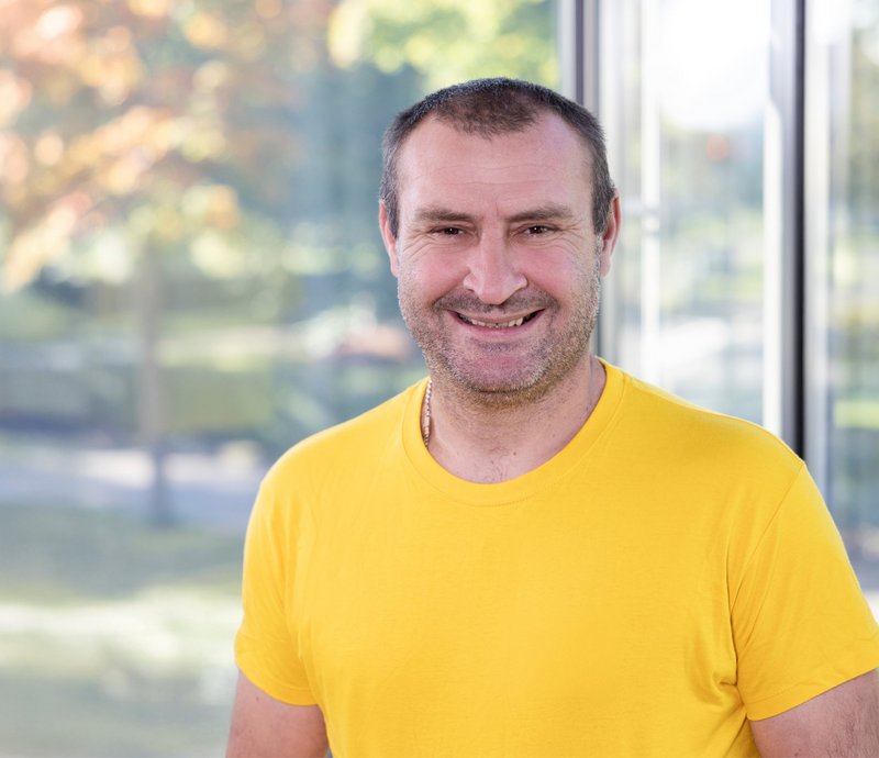 Regiebetrieb – Ihor Hrom Ein lächelnder Mann im gelben T-Shirt steht vor einem Fenster und schaut freundlich in die Kamera.