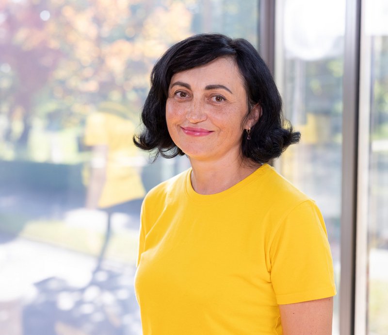 Olha Herasymenko Porträt einer lächelnden Frau mit schwarzen Haaren, die ein gelbes T-Shirt trägt, vor einem Fenster mit unscharfem Außenbereich.