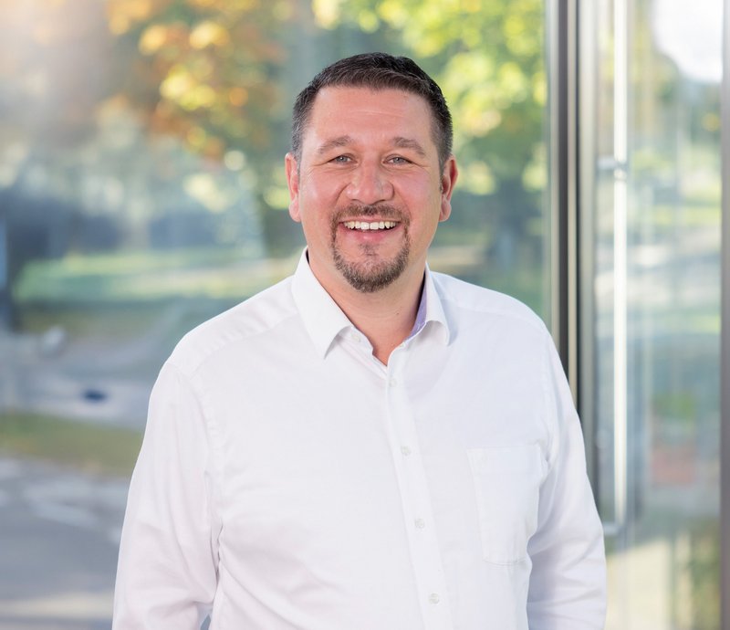 Michael Allweier SWG Friedrichshafen Porträt eines freundlich lächelnden Mannes in weißem Hemd vor einem modernen Gebäude, der direkt in die Kamera schaut.