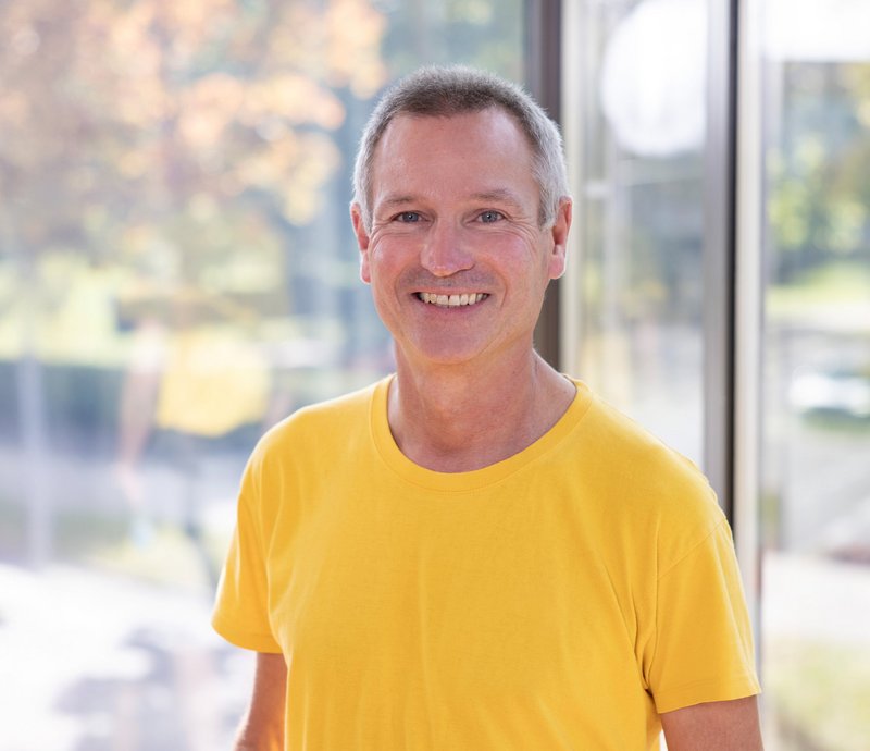 Karl-Heinz Höpfl Mittelalterlicher Mann in gelbem T-Shirt lächelt freundlich in die Kamera, im Hintergrund unscharf helle Außenumgebung.
