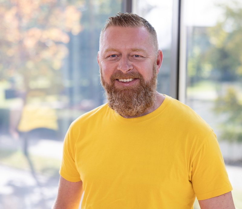 Elvir Osmanovic Ein lächelnder Mann mit kurzem, blonden Haar und Vollbart trägt ein gelbes T-Shirt. Er steht vor einem Fenster mit unscharfem, natürlichem Hintergrund aus Bäumen und Tageslicht.